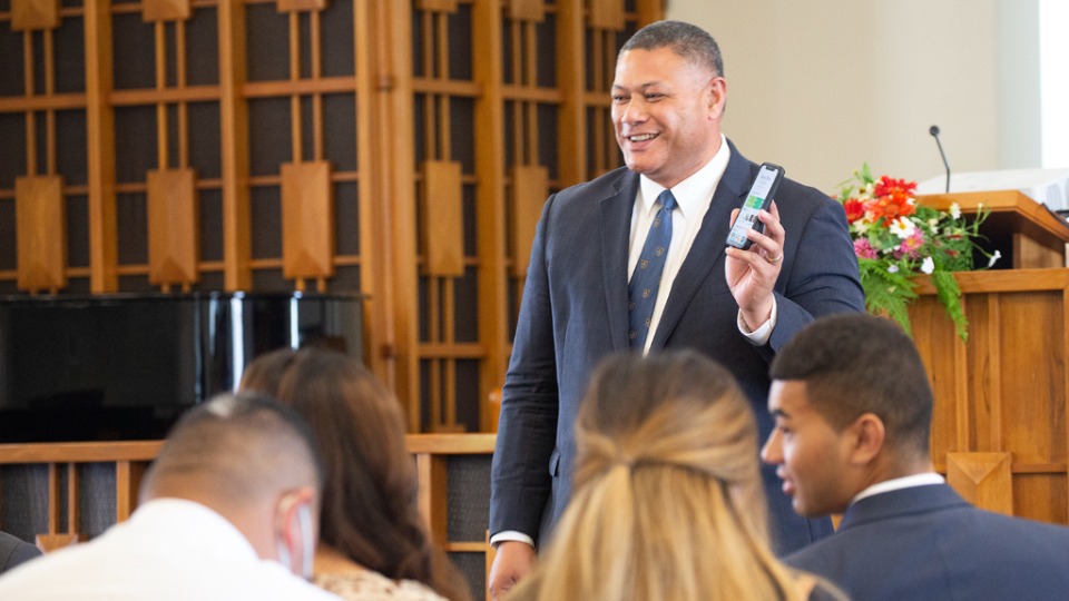 Bishop-of-the-Auckland-YSA-1st-Ward,-Sione-Tuione,-holds-up-his-phone-showing-the-Pacific-Area-app-as-he-discusses-the-Area-Presidency-Focus-for-2022-with-his-congregation.-